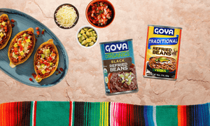 Various Goya refried beans with Cinco de Mayo theme background. Dice tomatoes, avocado and shredded cheese next to it. 