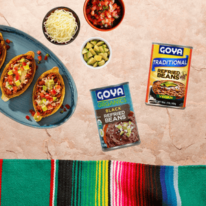 Various Goya refried beans with Cinco de Mayo theme background. Dice tomatoes, avocado and shredded cheese next to it. 