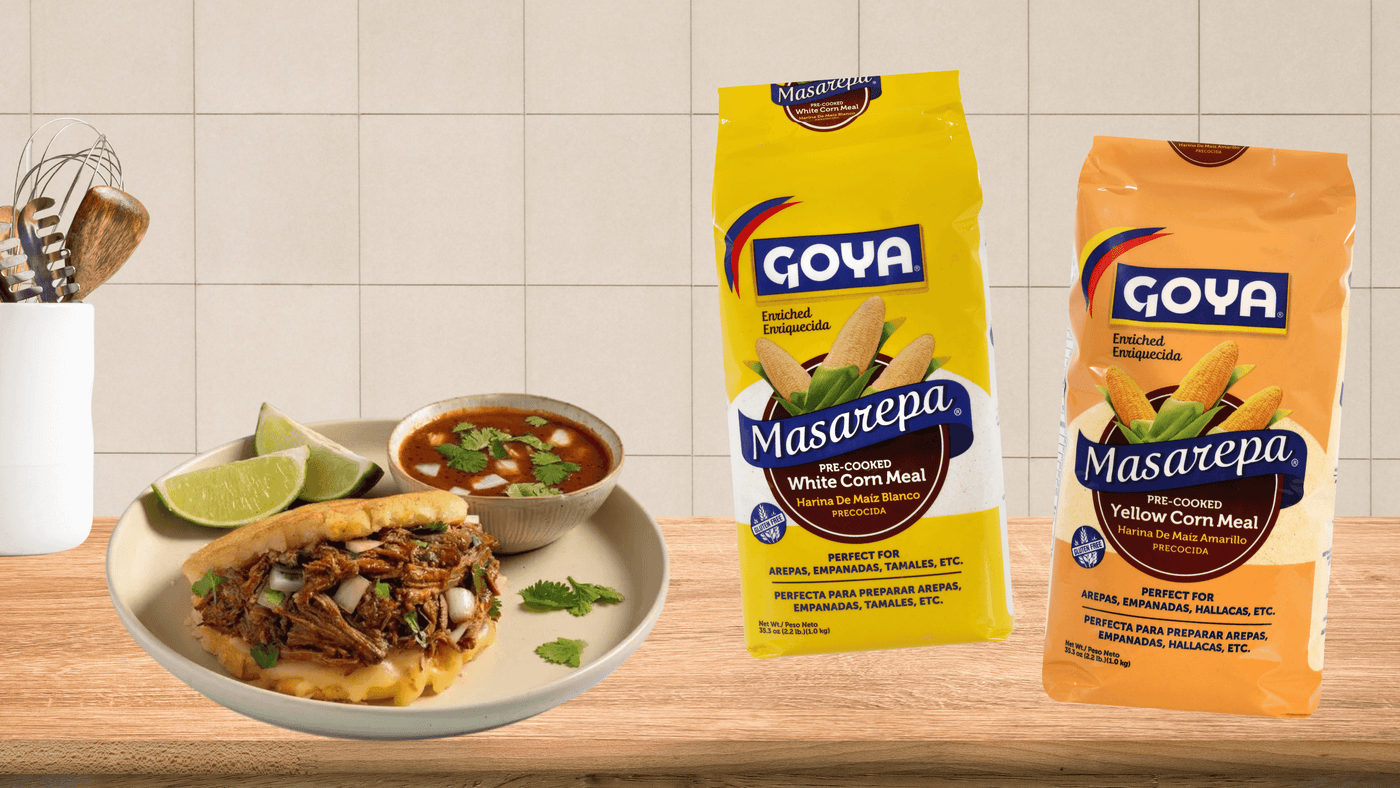 Plate with a savory arepa, lime wedges, and soup on a wooden table. Two Goya Masarepa cornmeal bags in the background. Cozy kitchen vibe.