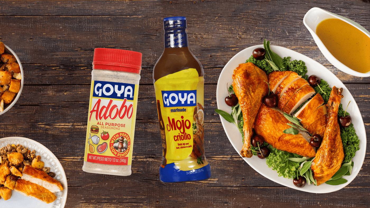 Goya Adobo and Mojo Criollo bottles are placed on a wooden table beside a platter of roasted chicken with greens and cherries. Additional dishes are visible on the side.