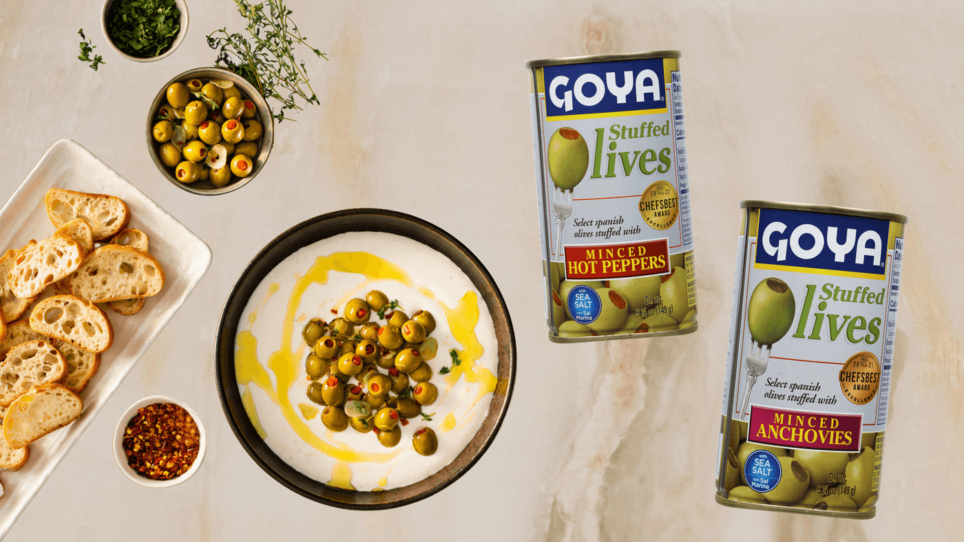 A bowl of olives in white sauce with olive oil drizzle is surrounded by sliced bread, herbs, and pepper flakes. Two Goya stuffed olives cans are displayed.